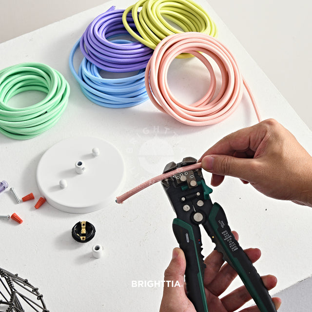 Making a white pendant light fixture with light color fabric cords on white surface. A hand holding a wire stripper stripping a pink fabric cord.